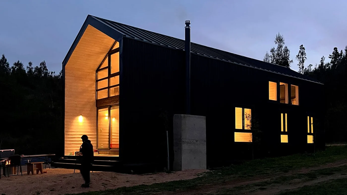Casa VG, Lago Vichuquén — volumen de envolvente negra al atardecer con la testera de madera iluminada como una linterna